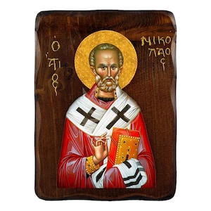 May include: A hand-painted icon of Saint Nicholas, also known as Santa Claus, on a wooden background. The icon depicts Saint Nicholas wearing a red and white robe with a gold cross and holding a book. The text "O TI" and "NIKO LAO" is written in gold on the wooden background.
