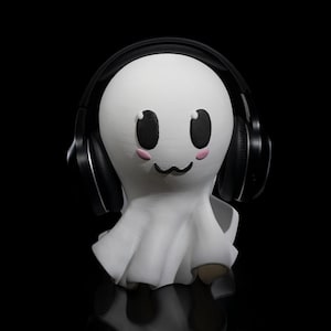 May include: A white ghost figurine with a cute face wearing black over-ear headphones. The ghost has pink cheeks and black eyes.