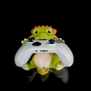 May include: A green and orange dinosaur-shaped controller stand holds a white Xbox controller. The dinosaur has a toothy grin and orange spikes on its head.