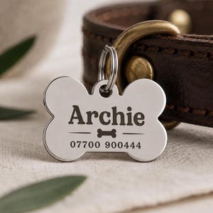 May include: A silver-toned bone-shaped pet identification tag attached to a dark brown leather collar. The tag is engraved with the name Archie, a small bone graphic, and the phone number 07700 900444 in a dark, clear sans-serif font.