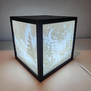 May include: A black cube-shaped lamp with a white frosted glass panel depicting a winter scene with trees, a crescent moon, and stars. The lamp is illuminated from within, casting a warm glow.