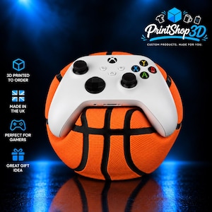 May include: A white wireless video game controller with black joysticks and coloured action buttons, resting on a textured orange and black basketball-shaped stand. The stand features a dimpled surface and black lines mimicking a basketball.