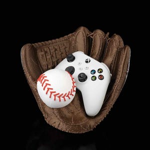 May include: A white Xbox controller and a white baseball are inside a brown leather baseball glove.