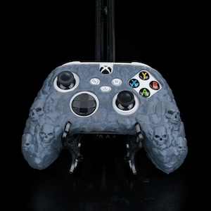 May include: A grey and black Xbox controller with a skull and bone design. The controller is on a stand.