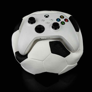 May include: A white Xbox controller is placed on top of a white football with black panels. The controller has black buttons and a black Xbox logo.