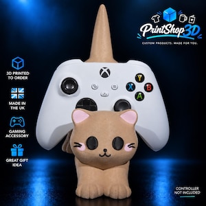 May include: A tan, cat-shaped 3D printed gaming controller stand featuring a pointed horn on top. The cat has black eyes, pink cheeks, and a small mouth. A white Xbox controller rests on the stand. Text includes PrintShop3D, 3D printed to order, made in the UK, gaming accessory, great gift idea, and controller not included.