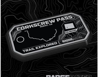 May include: A black and silver metal badge with the text "Corkscrew Pass, Ouray, CO" and "Trail Explored" with a map of the area and a small Colorado flag. The badge is labeled "BadgeRated".