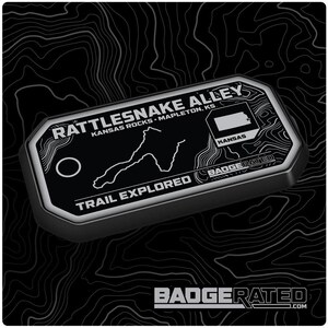 May include: A black and silver rectangular metal badge with the text "Rattlesnake Alley" and "Trail Explored". The badge features a map of Kansas and the text "Kansas Rocks - Mapleton, KS". The brand name "Badgerated" is also visible.