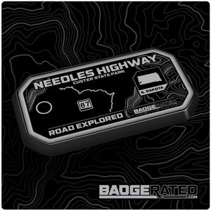 May include: A black and silver rectangular badge with the text "Needles Highway Custer State Park" and a map of South Dakota. The badge also includes the text "Road Explored" and the number "87". The brand name "Badgerated" is also visible.