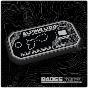 May include: A black and silver metal badge with the text "ALPINE LOOP OURAY LAKE CITY SILVERTON, CO" and "TRAIL EXPLORED" along with a map of the area and the state of Colorado flag. The badge is labeled "BADGERATED" and has a small hole at the top for attaching it to something.