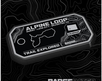 May include: A black and silver metal badge with the text "ALPINE LOOP OURAY LAKE CITY SILVERTON, CO" and "TRAIL EXPLORED" along with a map of the area and the state of Colorado flag. The badge is labeled "BADGERATED" and has a small hole at the top for attaching it to something.