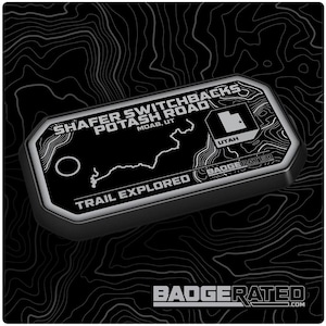 May include: Black and white metal badge with the text "Shafer Switchbacks Potash Road Moab, UT" and a map of the trail. The badge also includes the text "Trail Explored" and "BadgeRated".