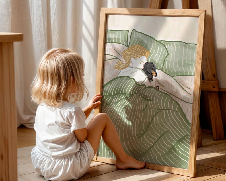 May include: A framed art print featuring a whimsical illustration of a person and a dachshund dog sleeping under green and white striped bedding. The artwork is in a wooden frame. A young child is sitting on the floor, looking at the artwork.