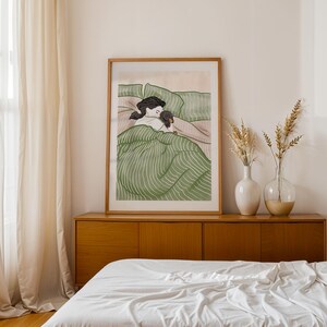 May include: A framed art print depicts a person and a dog in bed, under a green striped blanket. The artwork is in a wooden frame and is displayed above a wooden cabinet. Two vases with dried plants are on the cabinet.