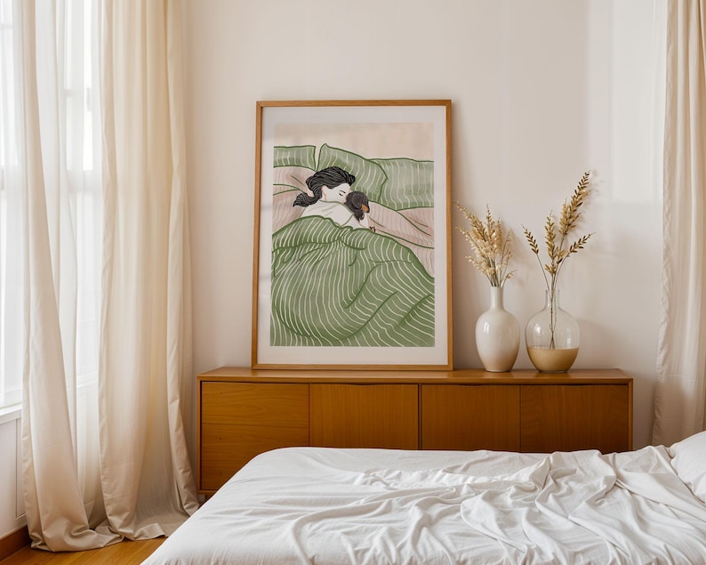 May include: A framed art print depicts a person and a dog in bed, under a green striped blanket. The artwork is in a wooden frame and is displayed above a wooden cabinet. Two vases with dried plants are on the cabinet.