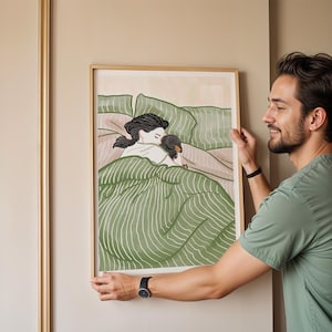 May include: A framed art print depicts a person and a dachshund dog snuggled in bed. The artwork features a green and white striped blanket, with the person's hair in a ponytail. The frame is a light wood color.