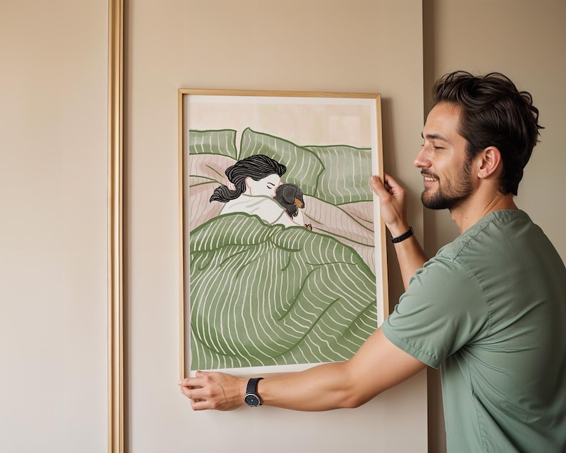 May include: A framed art print depicts a person and a dachshund dog snuggled in bed. The artwork features a green and white striped blanket, with the person's hair in a ponytail. The frame is a light wood color.