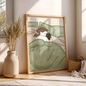 May include: A framed art print depicts a person and a dog in bed. The illustration features a person with brown hair and a black and brown dog nestled under green striped bedding. The artwork is in a light wooden frame.