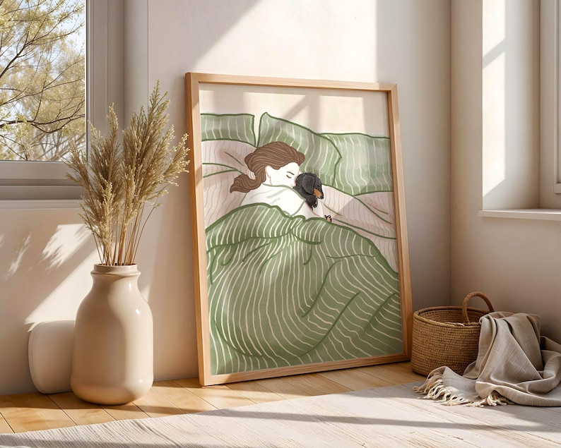 May include: A framed art print depicts a person and a dog in bed. The illustration features a person with brown hair and a black and brown dog nestled under green striped bedding. The artwork is in a light wooden frame.
