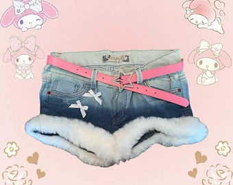 Può includere: Pantaloncini di jeans con cintura rosa e fibbia a forma di stella. Gli shorts hanno finiture in pelliccia bianca e piccoli fiocchi bianchi. Personaggi dei cartoni animati e la parola "MEOW" sullo sfondo.