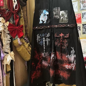 Puede incluir: Falda de mezclilla negra con diseños gráficos rojos, blancos y azules. La falda tiene varios bolsillos y un detalle de cadena.