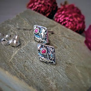May include: A pair of silver stud earrings with a textured design and a small, round, red gemstone in the center. The earrings are square-shaped with a detailed, organic pattern. Earring backs are visible. The background includes pink flowers.