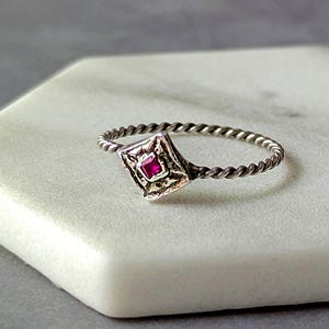 May include: A silver ring with a twisted band and a square, ornate setting. The setting holds a small, square, ruby-colored stone. The ring is displayed on a white, hexagonal surface against a gray background.