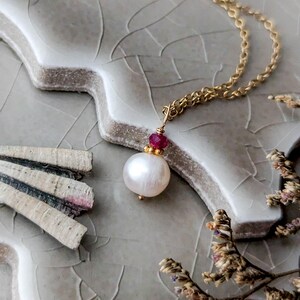 May include: A delicate gold necklace featuring a large, luminous white pearl pendant. The pendant is accented with a small, vibrant red gemstone and suspended from a fine gold chain. The necklace is displayed on a textured, gray surface.