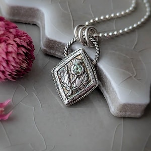 May include: A silver pendant necklace with a diamond-shaped pendant. The pendant features a thistle design with a small green gemstone in the center. The necklace is on a silver chain, and a pink flower is in the background.