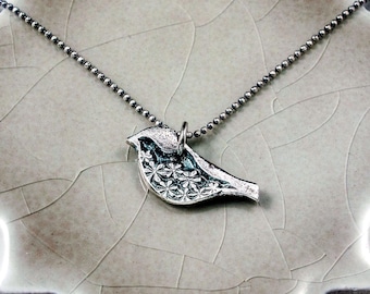May include: A silver-toned bird pendant necklace. The bird pendant features a detailed floral pattern on its body and hangs from a delicate beaded chain. The necklace is displayed on a light gray, textured surface.