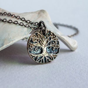 May include: A silver-toned pendant necklace featuring a detailed tree of life design. The round pendant has an intricate, textured pattern with a dark background. The necklace chain is also silver-toned, and the pendant is displayed on a light-colored surface.