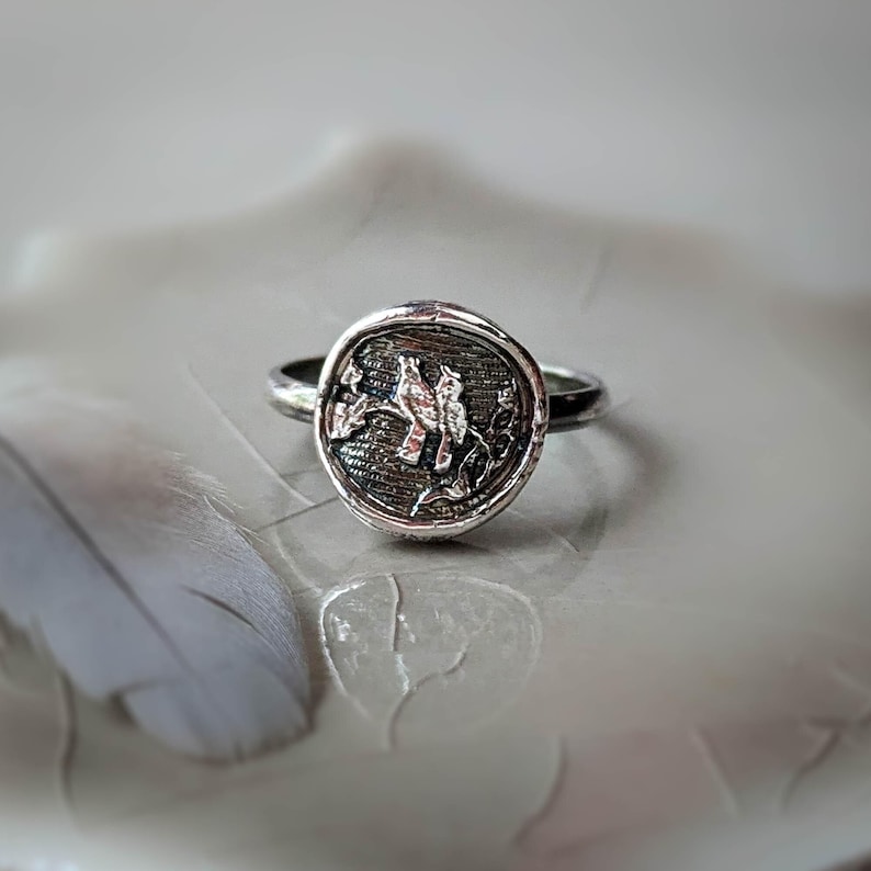 May include: A silver ring with a round, textured design featuring two birds perched on a branch. The ring is on a white surface.