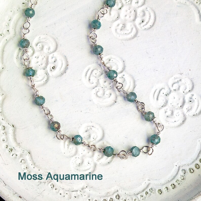 May include: A delicate silver chain necklace with moss aquamarine beads. The necklace is on a white background with a floral pattern.