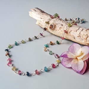 Puede incluir: Un delicado collar con pequeñas piedras preciosas de colores ensartadas en una cadena de plata. Las piedras preciosas son de varios colores, incluyendo rosa, verde, azul y amarillo. El collar se exhibe sobre un trozo de madera flotante, con una flor morada en el fondo.
