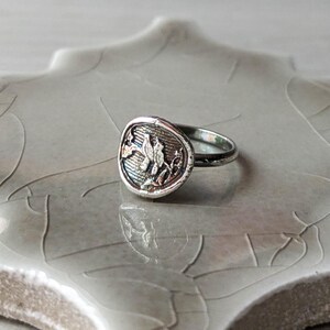 May include: A silver ring with a round, textured top featuring a detailed floral design. The ring is on a light gray background.