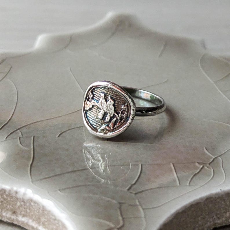 May include: A silver ring with a round, textured top featuring a detailed floral design. The ring is on a light gray background.