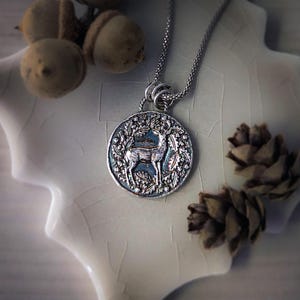 May include: Silver necklace with a round pendant featuring a deer standing in a wreath of leaves. The pendant has a textured, antique finish.