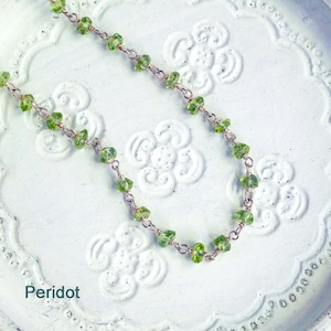 May include: A delicate necklace made with small, green peridot stones and silver chain. The necklace is displayed on a white background with a floral pattern.