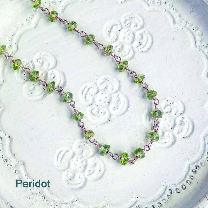 May include: A delicate necklace made with small, green peridot stones and silver chain. The necklace is displayed on a white background with a floral pattern.