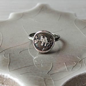 May include: A silver ring with a round, textured bezel featuring a detailed design of two birds perched on a branch. The ring is on a white, textured surface.