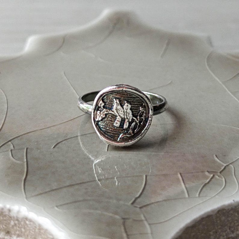 May include: A silver ring with a round, textured bezel featuring a detailed design of two birds perched on a branch. The ring is on a white, textured surface.