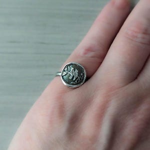 May include: A silver ring with a round, textured, green and silver design. The design features a stylized floral motif.