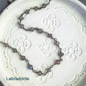 May include: A necklace made with labradorite stones and wire. The stones are a dark gray with blue and green iridescent flashes. The necklace is on a white background with a floral pattern.