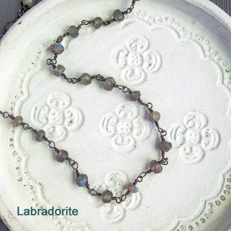 May include: A necklace made with labradorite stones and wire. The stones are a dark gray with blue and green iridescent flashes. The necklace is on a white background with a floral pattern.