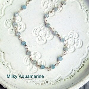 May include: A delicate silver chain necklace with light blue aquamarine beads. The necklace is on a white background with a floral pattern. Milky Aquamarine.