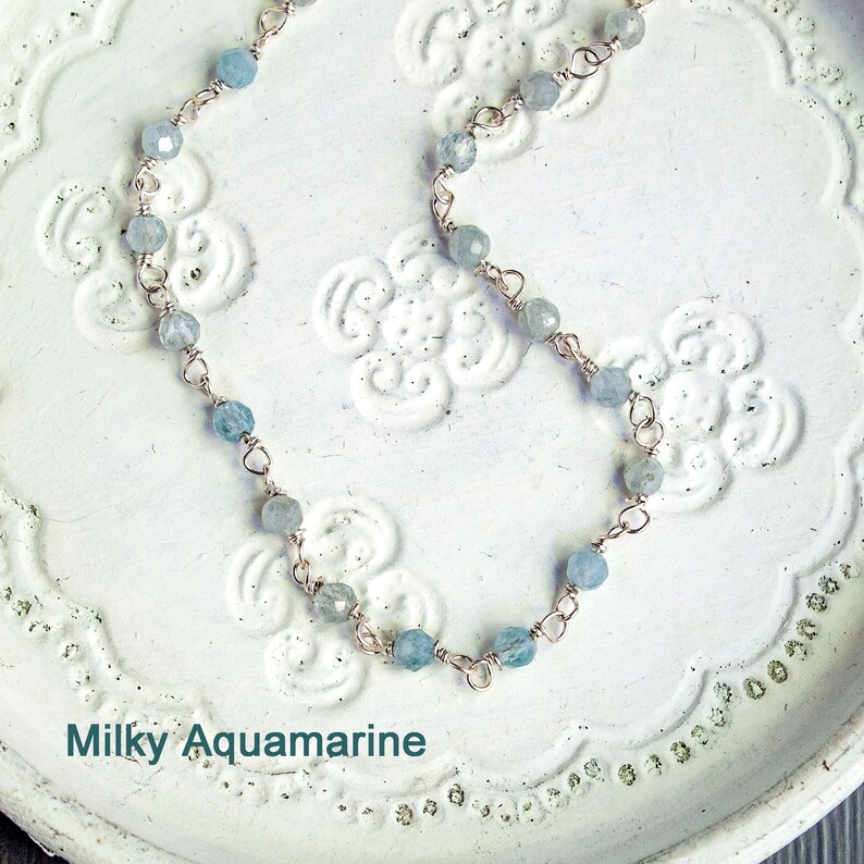 May include: A delicate silver chain necklace with light blue aquamarine beads. The necklace is on a white background with a floral pattern. Milky Aquamarine.