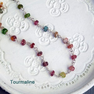 May include: A silver chain necklace with multi-colored tourmaline gemstones. The necklace is on a white background with a floral pattern. Tourmaline.