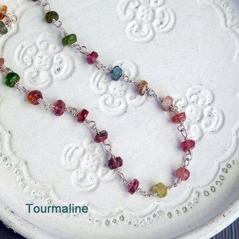 May include: A silver chain necklace with multi-colored tourmaline gemstones. The necklace is on a white background with a floral pattern. Tourmaline.