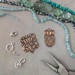 May include: A selection of jewellery-making components. The image shows strands of beads in shades of blue, green, and black. There are also metal clasps, decorative metal pendants, and a pair of pliers. The pendants feature detailed floral patterns.