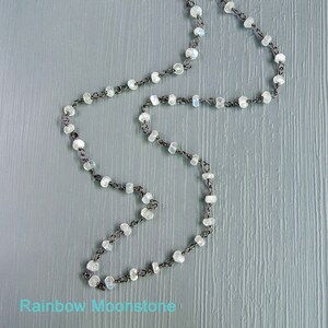 May include: A silver chain necklace with rainbow moonstone beads. The necklace is on a light gray wooden surface.